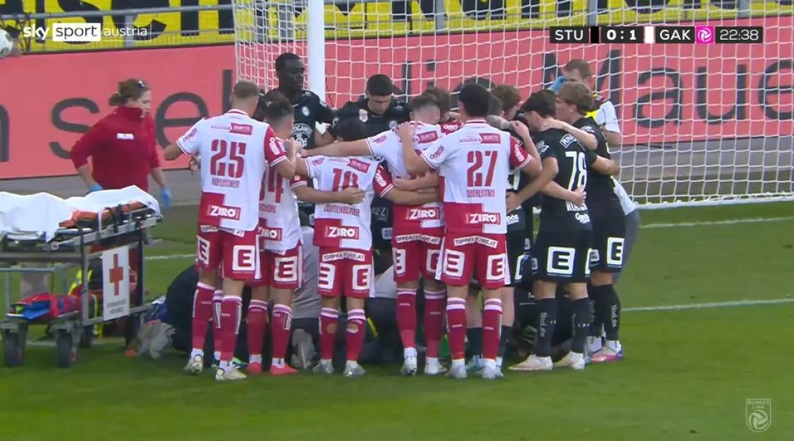 Players from both teams huddle around a goalkeeper who suffered a horrific injury on the pitch, with medical staff and a stretcher nearby.