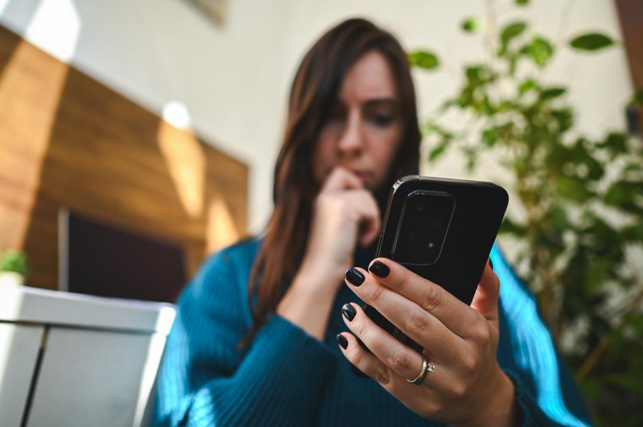 A woman in a blue sweater looks worried as she holds a black smartphone in her hands, contemplating a call from an unknown number.