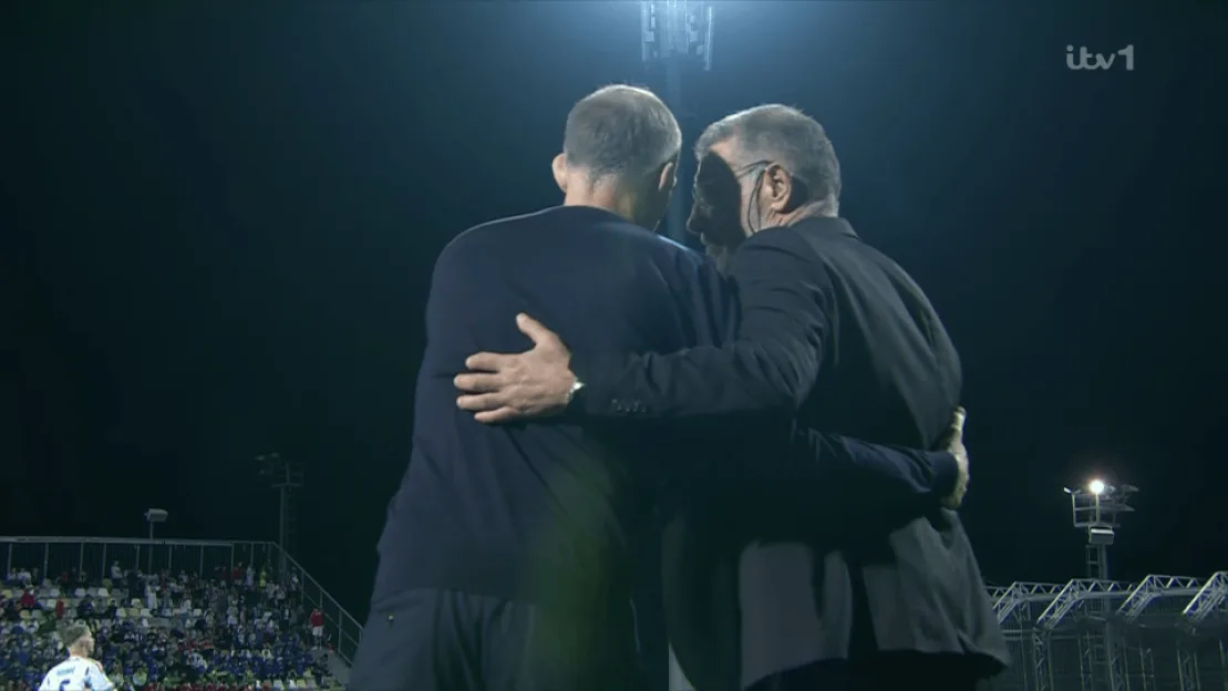 Two men embrace on a football pitch at night under floodlights.