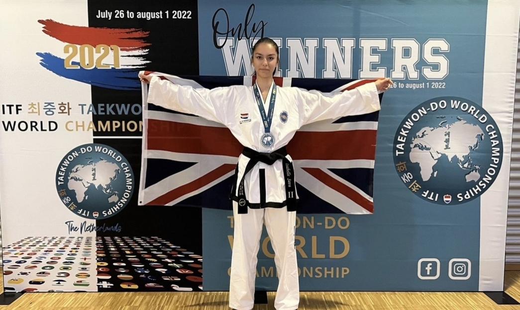 A young woman in a taekwondo uniform holding up a British flag, standing in front of a backdrop for the ITF Taekwondo World Championships.