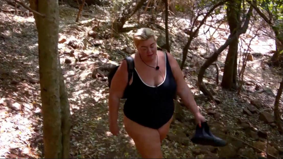 Gemma Collins wearing a black swimsuit while walking through a wooded area.