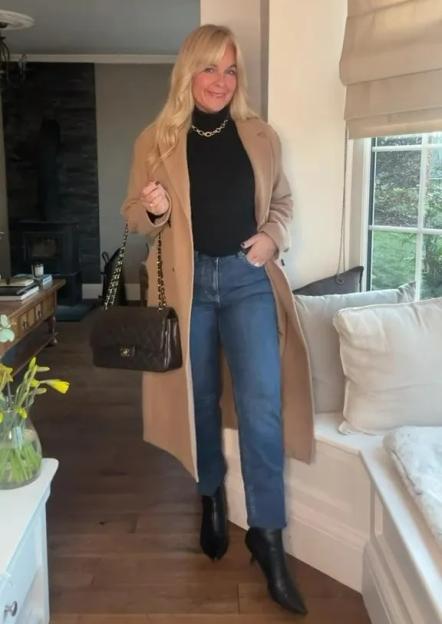 A woman wearing a tan coat, black turtleneck, blue jeans, and black boots, holding a quilted black handbag.