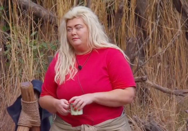 Gemma Collins with long blonde hair, wearing a red t-shirt, light brown skirt, and holding a small cup, against a backdrop of tall brown grass.
