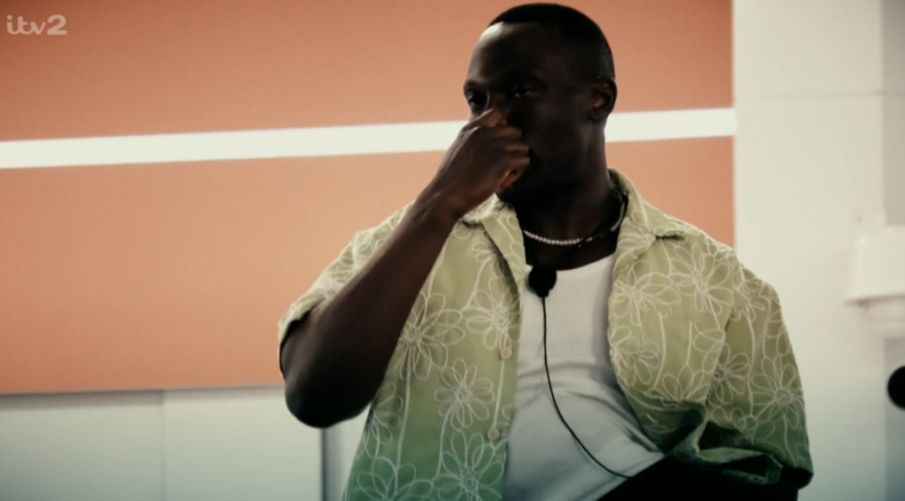 A dark-skinned man in a light green floral shirt pinches his nose.