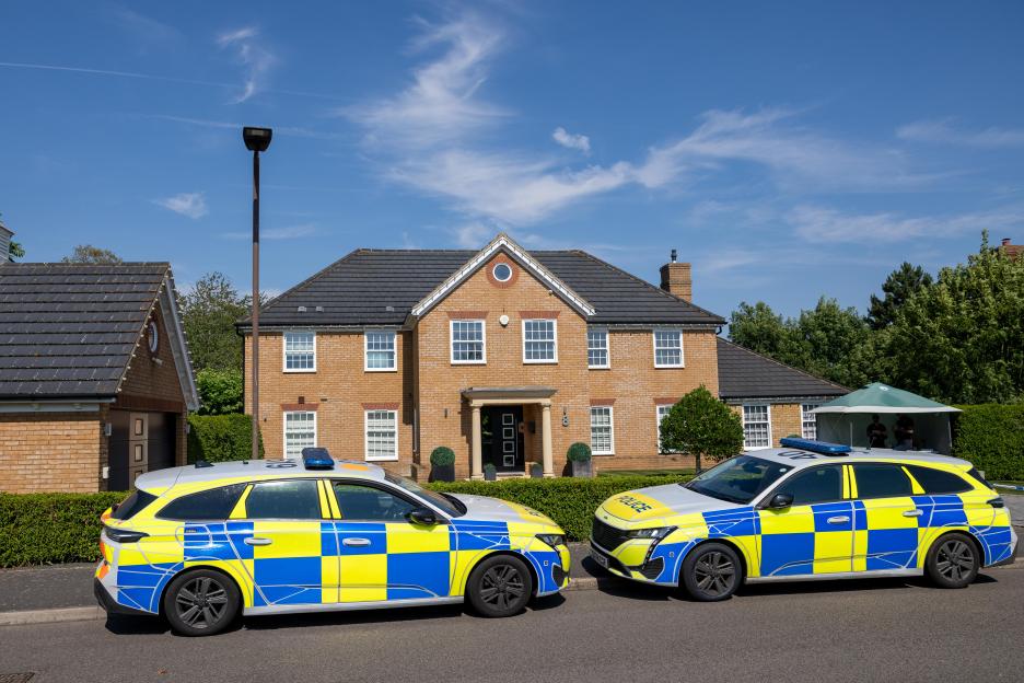 Two police cars parked outside a large brick house with a green tent set up on the lawn.