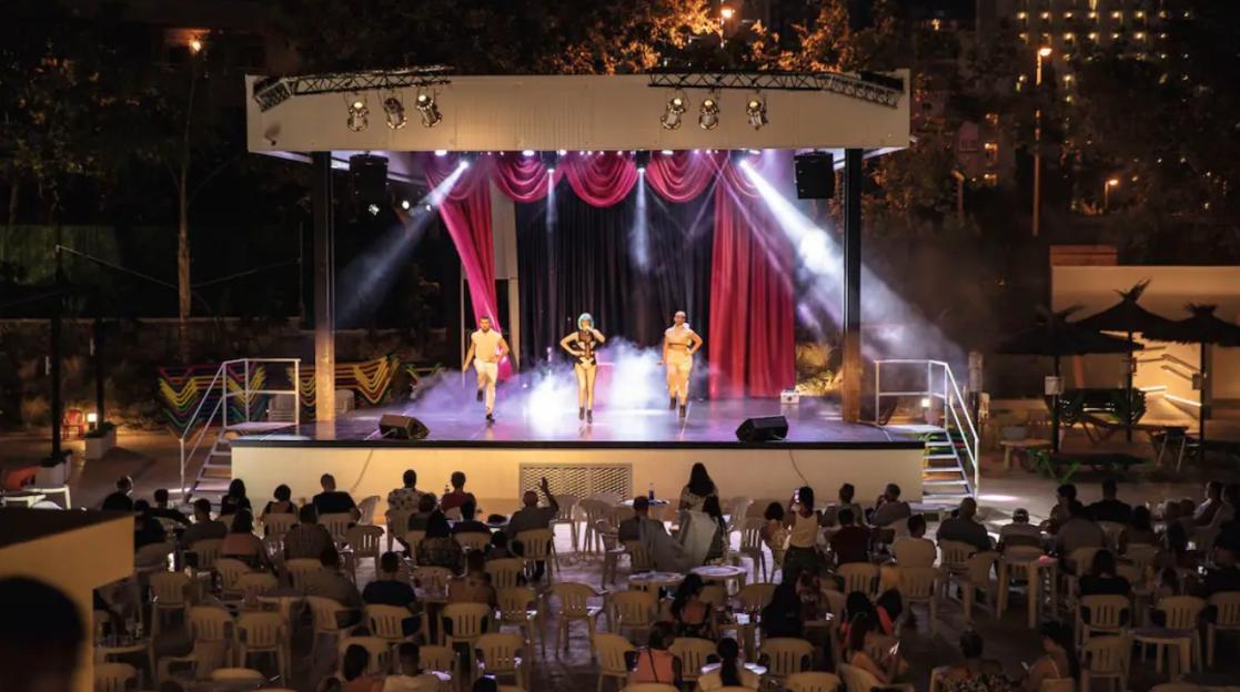 Three performers on a lit stage with an audience watching in front of it.