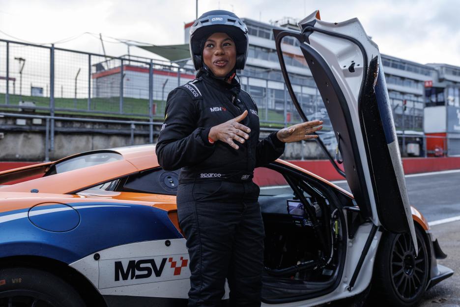 Kemi Badenoch, in a racing suit and helmet, reacts after a ride in a McLaren race car.