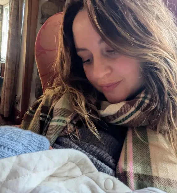 A woman with long brown hair, wearing a plaid scarf, gazes down with a gentle smile at a baby dressed in a light blue hat and quilted blanket.