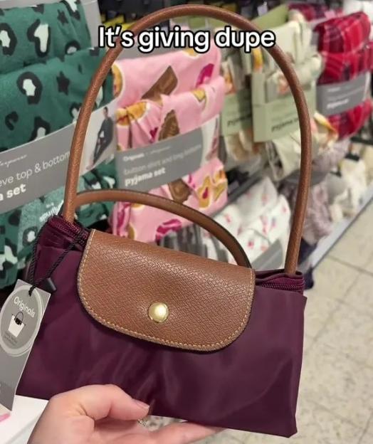 Hand holding a maroon foldable tote bag with a tan leather flap and handle, with pajamas displayed in the background.