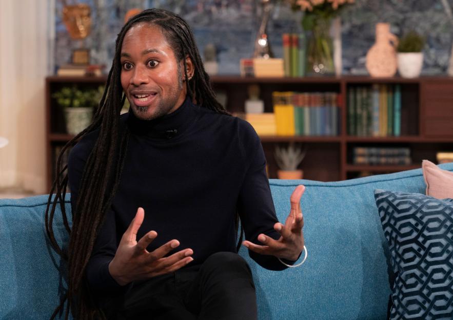 Professor Jason Arday, a Black man with dreadlocks, sitting on a blue couch and gesturing with his hands.