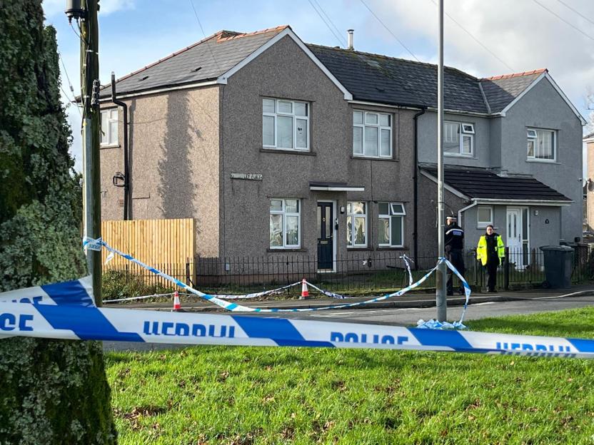 Police tape across a residential street with two officers standing near a house in the background.