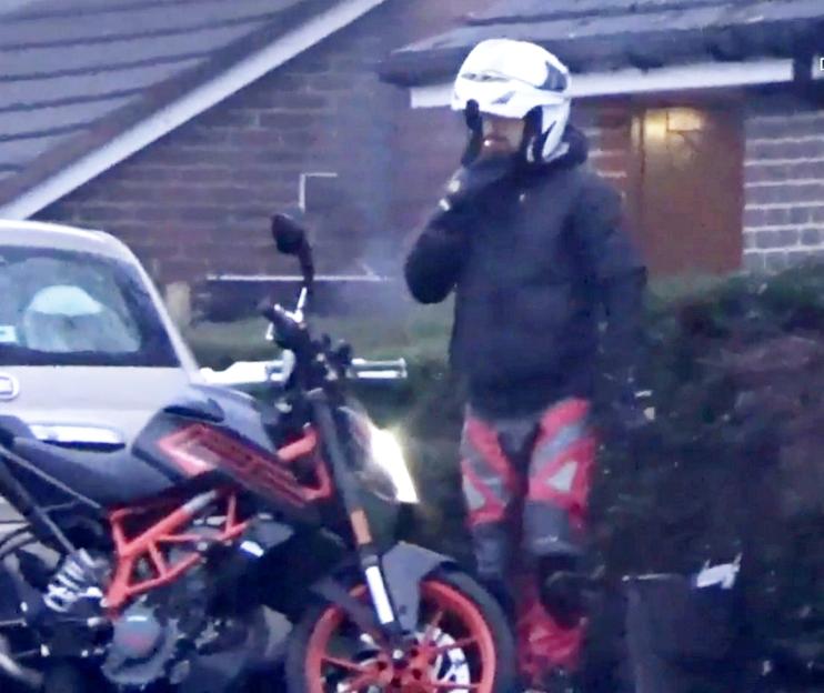 A man in a helmet, black jacket, and red and grey pants stands next to a motorcycle, smoking a cigarette.