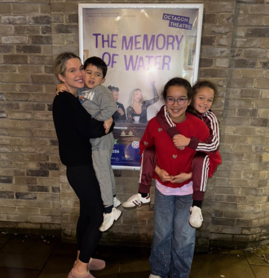 A woman holding a boy, with two girls piggybacking each other, standing in front of an Octagon Theatre poster for "The Memory of Water."