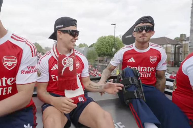 Two men in Arsenal football jerseys, one with a red and white scarf, on a bus during a parade.