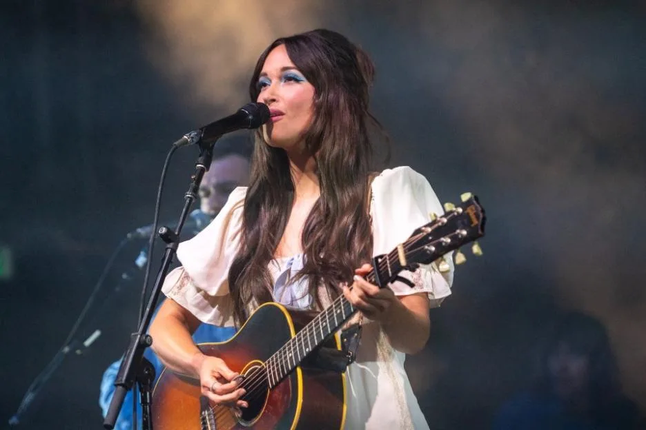 Kacey Musgraves singing and playing an acoustic guitar on stage.