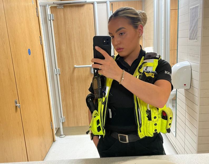 Rebecca Nicholls, a former police officer, wearing a high-visibility vest and black uniform, taking a selfie in a mirror.