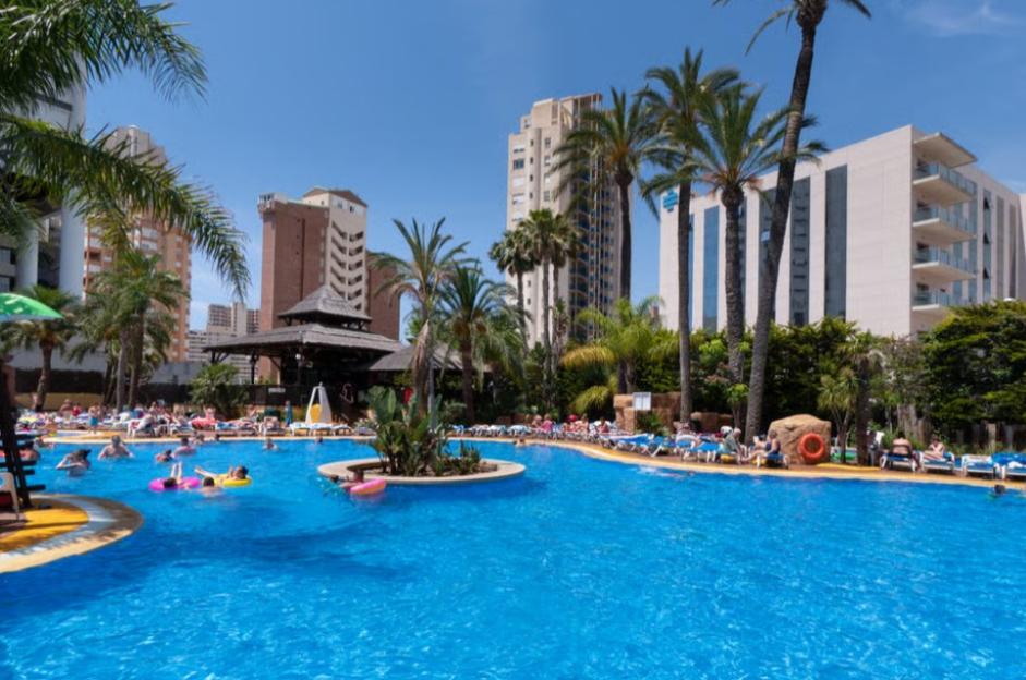 People relaxing in a large outdoor swimming pool area with palm trees and tall buildings in the background.