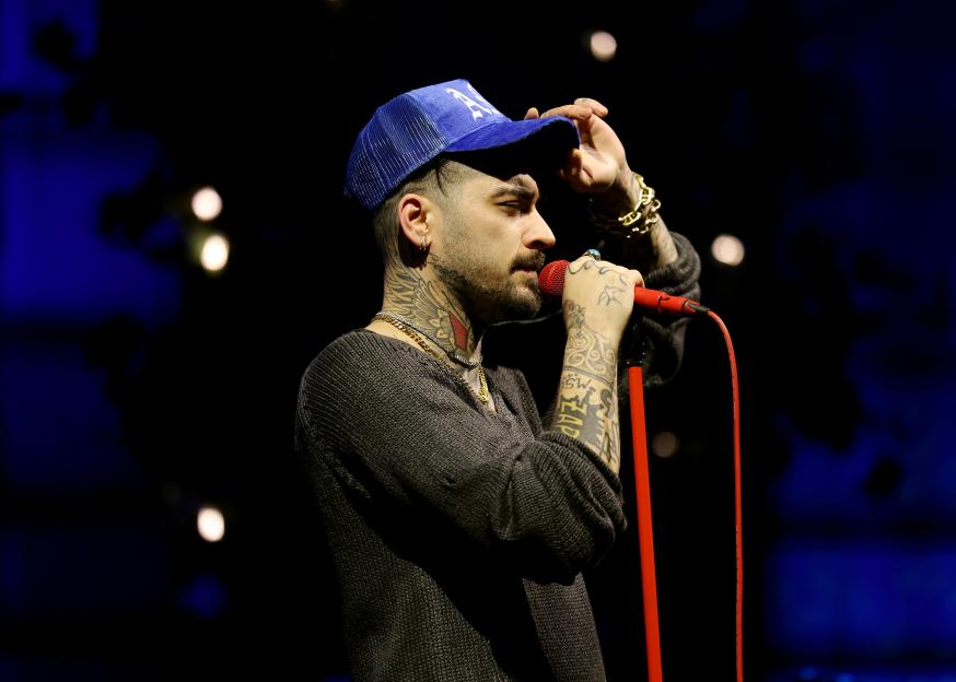 Zayn Malik performing on stage in a blue cap, dark grey sweater, and tattoos.
