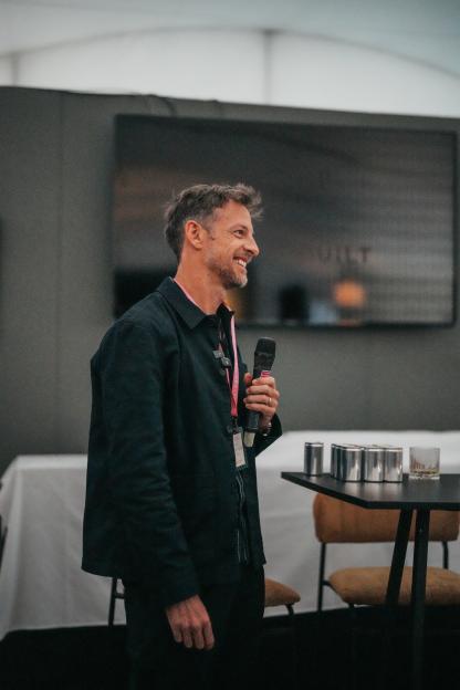 A smiling man with short brown hair and a beard, wearing a dark jacket, holding a microphone.