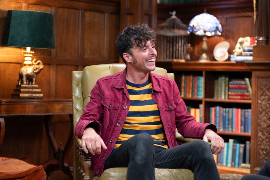 A man with curly hair, wearing a striped yellow and blue sweater and a red jacket, sitting in a leather armchair and smiling.