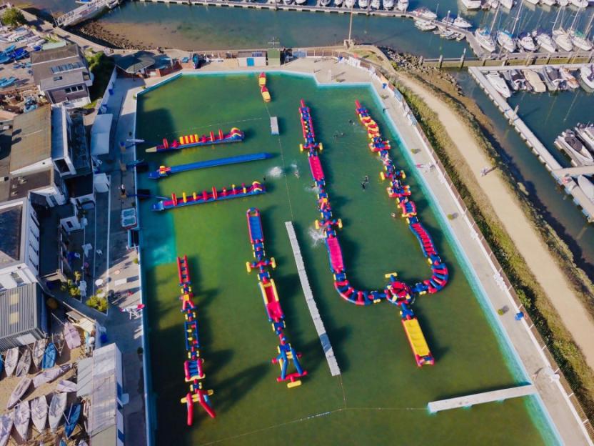 Aerial view of a water park with inflatable obstacles in a large pool next to a marina filled with boats.