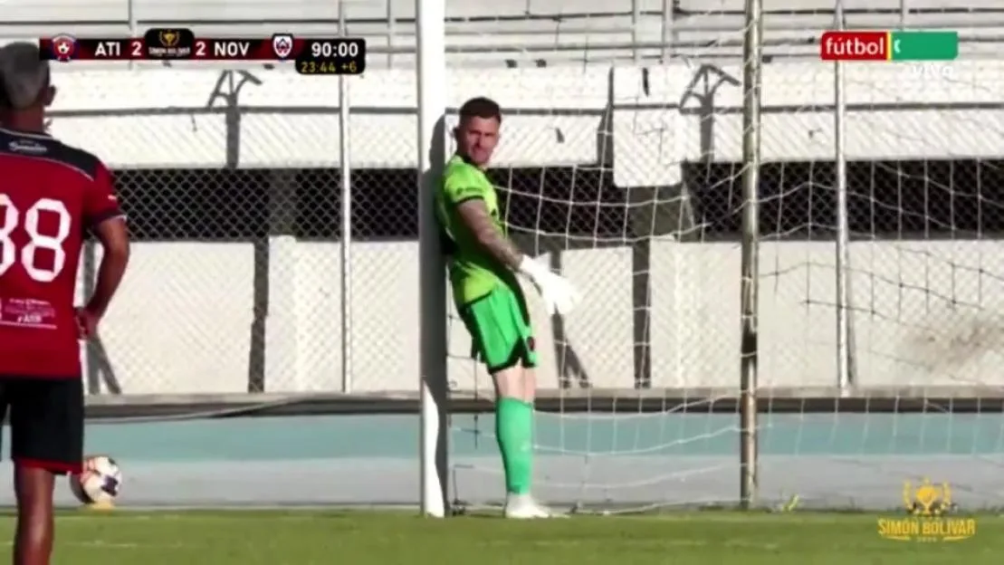 A soccer player in a green uniform and white gloves stands by a goalpost, looking toward the camera, with another player in a red jersey to the left.