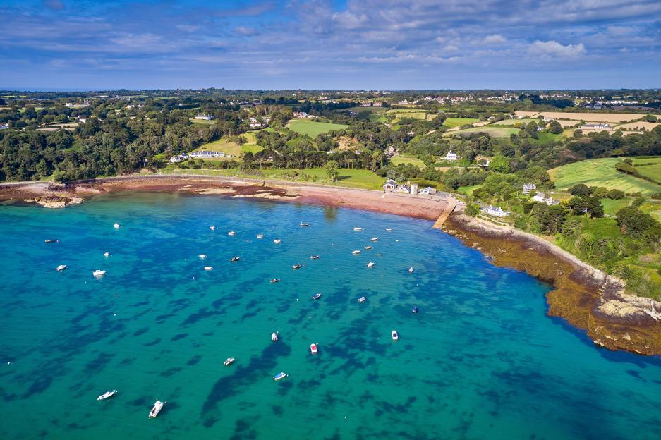 Aerial drone image of St Catherines Bay in Jersey CI, with green land, trees, and small buildings bordering the bay where many boats are anchored.