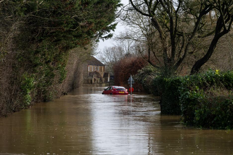 A red car partially submerged in brown floodwaters on a rural road in Martock, Somerset.