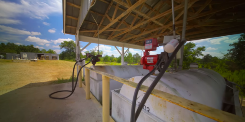 Fueling station with two large white tanks, a red fuel pump, and hoses under a wooden-beamed shed.