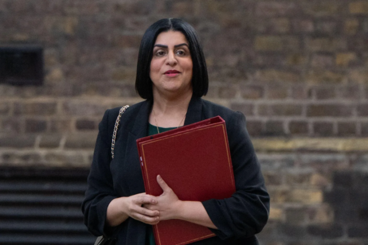 A woman with dark hair, a dark suit jacket, and a dark green shirt holding a red folder.