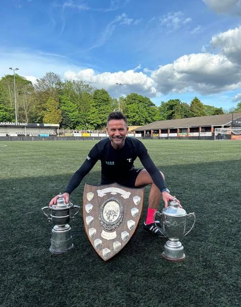 A smiling man on a soccer field holding two trophies and a shield.