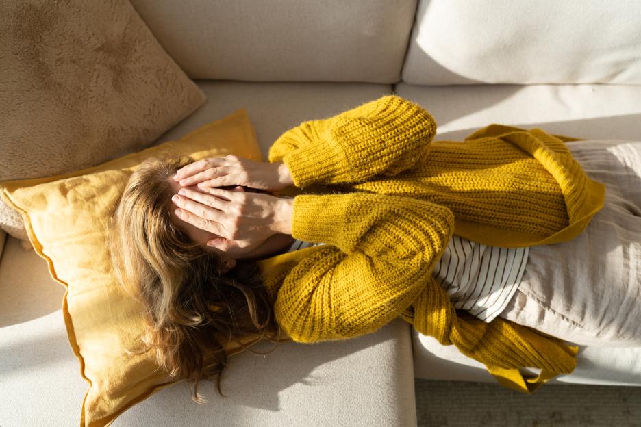 Woman lying on a sofa with hands covering her face, depicting stress or illness.