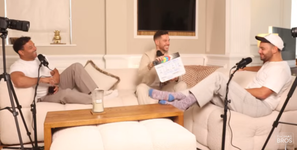 Three men sitting on a couch for a podcast, one holding a clapperboard.