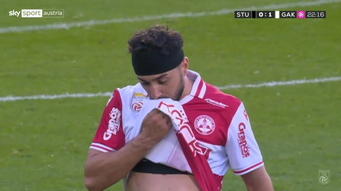 A soccer player wearing a red and white jersey pulls his shirt up over his face in distress on the pitch.