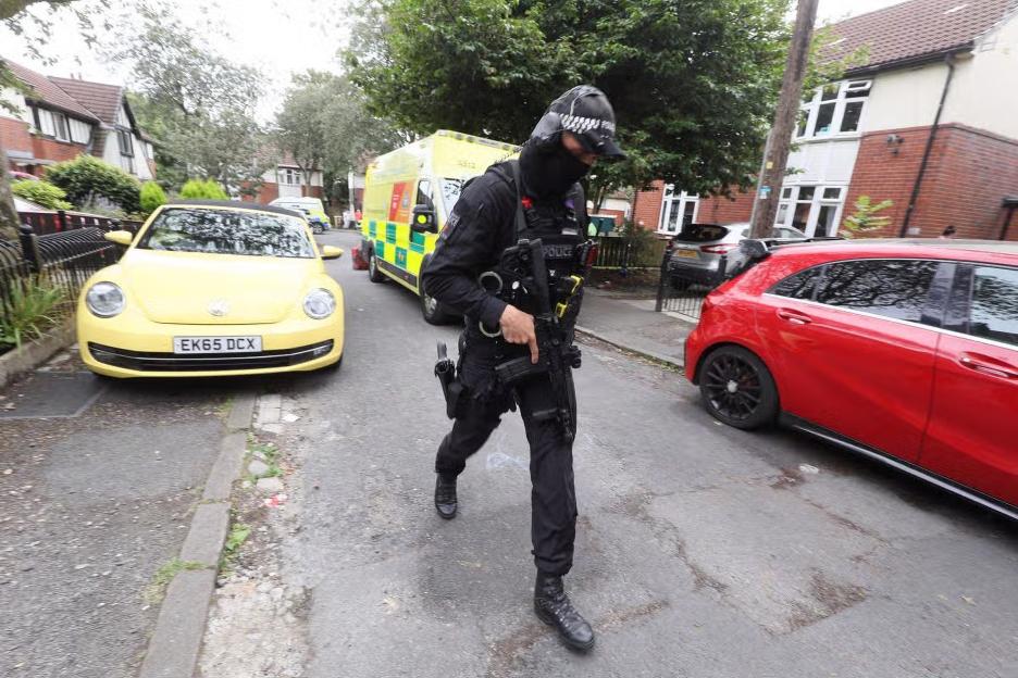 Armed police officer on a residential street with an ambulance in the background.