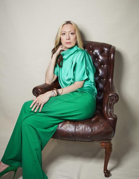Jen Barton Packer in a green blouse and pants, sitting in a leather armchair.