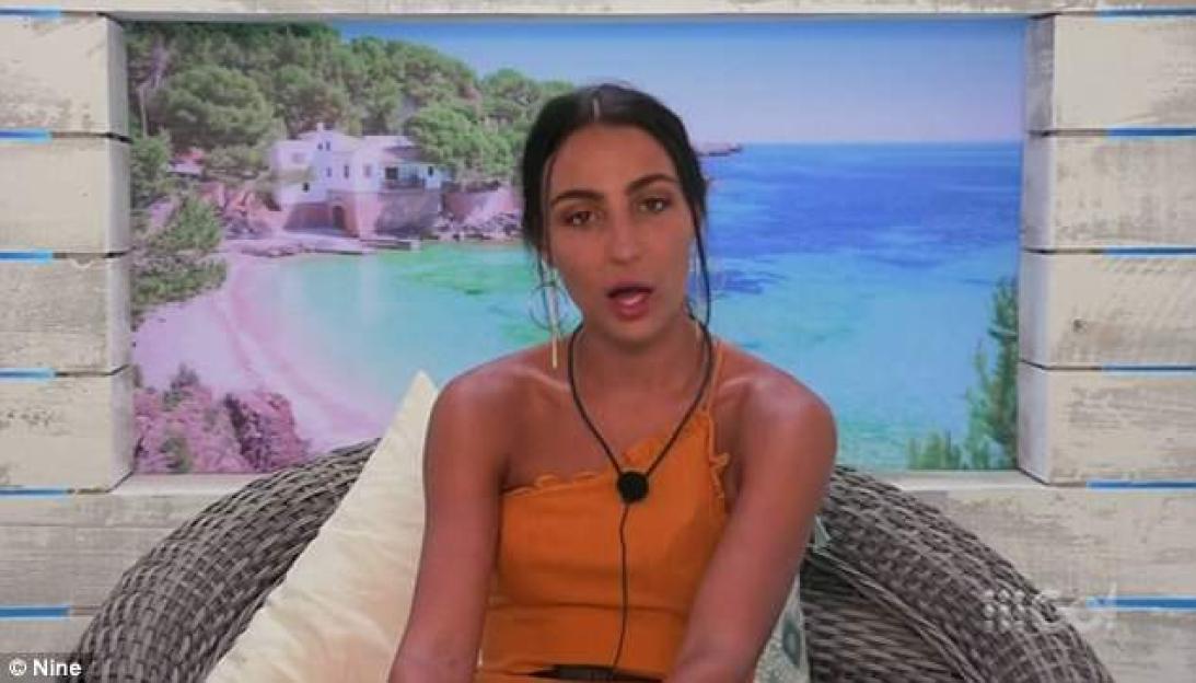 Taylor Broad from Love Island Australia, wearing an orange top and seated in a wicker chair, against a backdrop of a beach and ocean.