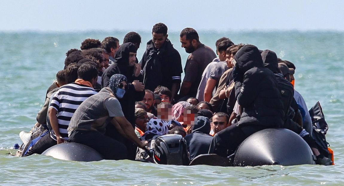 An overloaded migrant dinghy in the sea, packed with many men, women, and children.