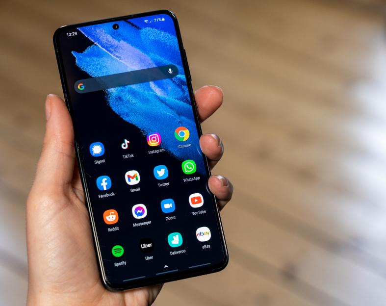 A hand holding a smartphone displaying various app icons on a dark blue background.