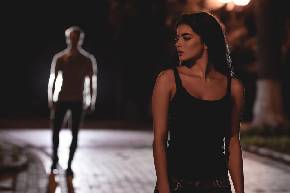 A woman looking concerned on a dark street with the blurred silhouette of a man in the background.