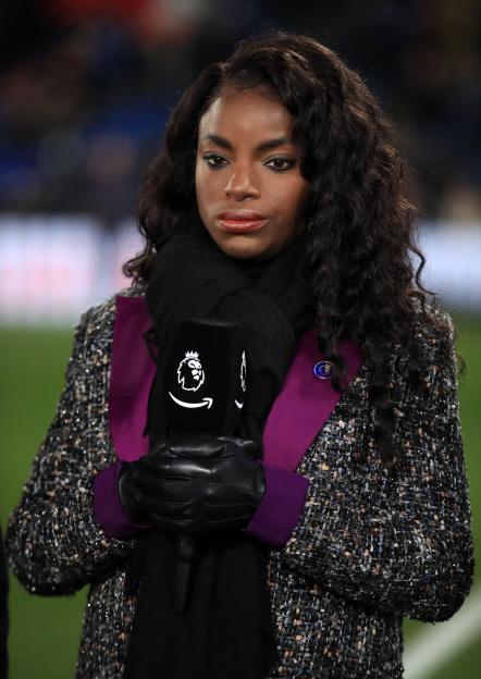 Eni Aluko, wearing a patterned coat, scarf, and gloves, holding a microphone.