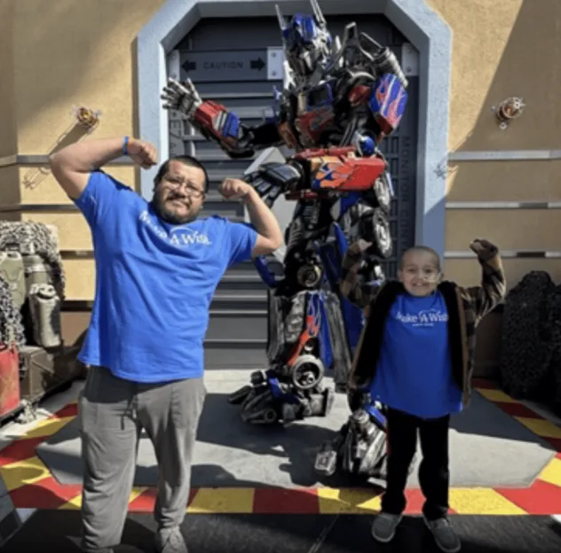 A man and a boy in Make-A-Wish shirts posing with Optimus Prime.