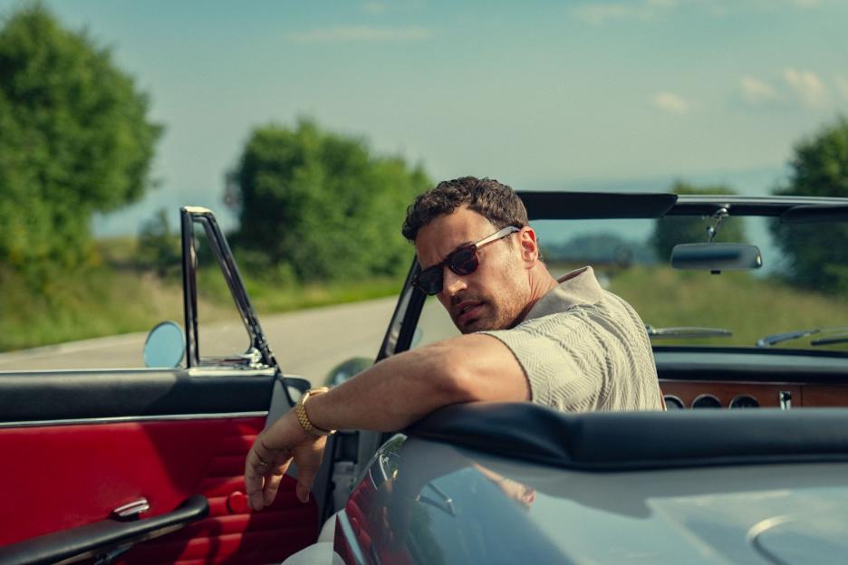 A man with sunglasses and a patterned shirt leans back in a convertible car, looking over his shoulder.