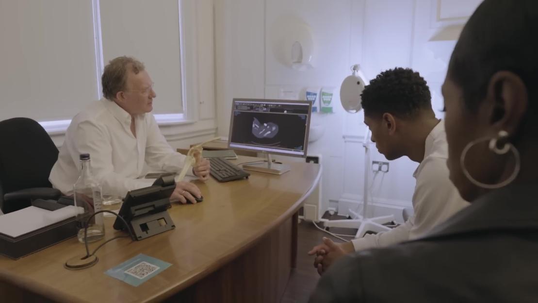 A doctor explaining an injury using a shoulder model and a scan on a computer screen to a young man and woman.