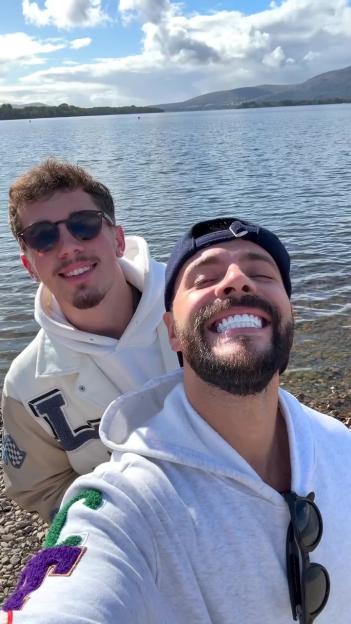 A man with a beard and a baseball cap smiles broadly with his eyes closed, next to a man with sunglasses, in front of a lake.