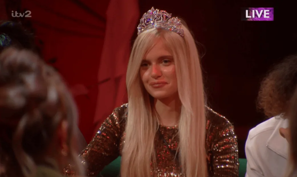 A woman with long blonde hair and a tiara wearing a sparkly bronze top sitting among other people, with ITV2 and LIVE logos visible in the corners.