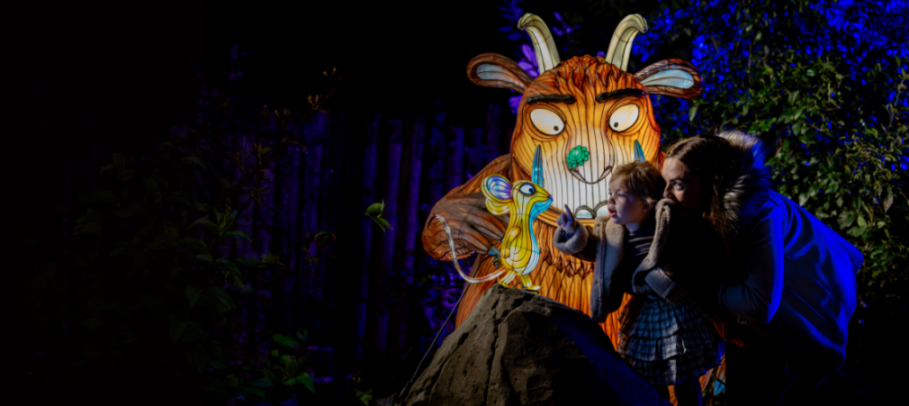 A child points at an illuminated mouse next to a large illuminated Gruffalo, with an adult observing at Twycross Zoo's Christmas lights.