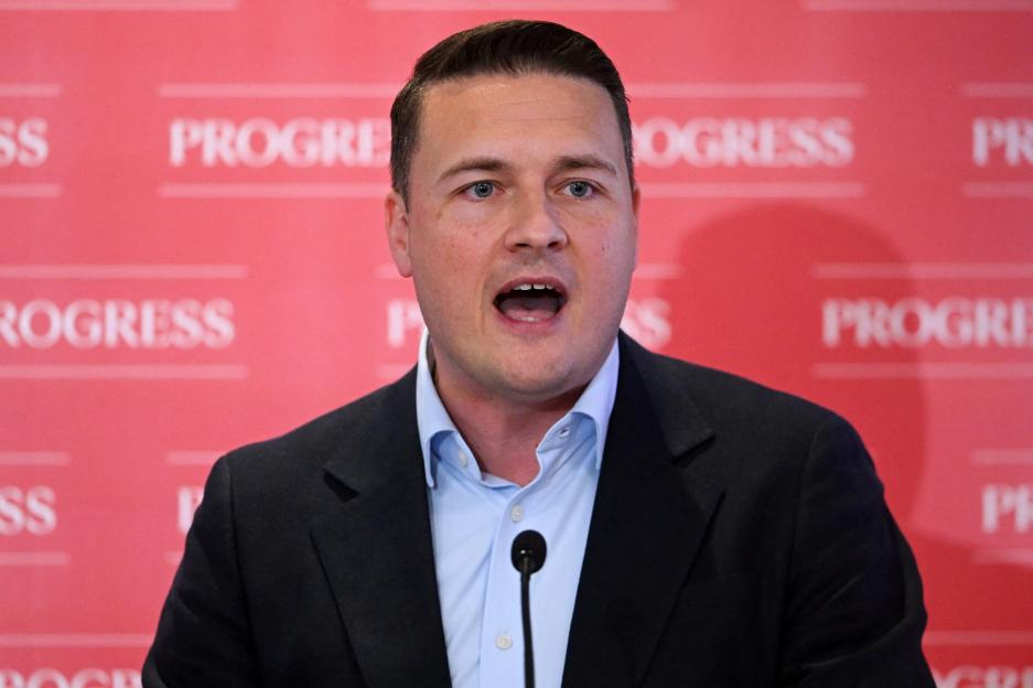 A man speaking into a microphone in front of a red backdrop with "PROGRESS" written repeatedly.
