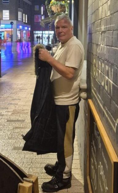 A man standing outside at night, holding a dark coat, with a blurry street and buildings in the background.