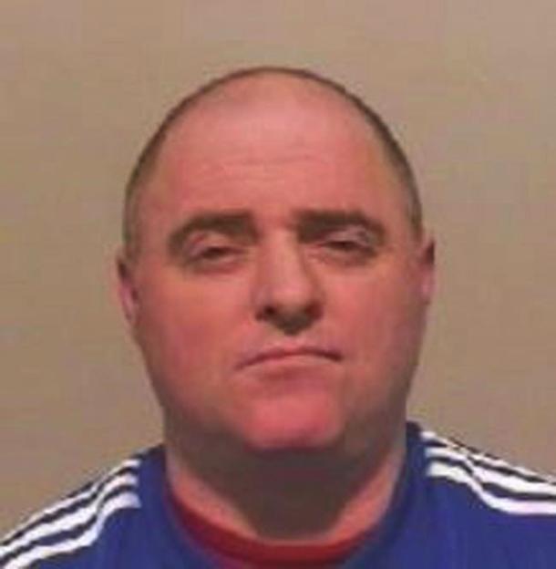 Mugshot of Raymond McDonald, a bald man with a serious expression, wearing a blue and red shirt with white stripes on the shoulders.